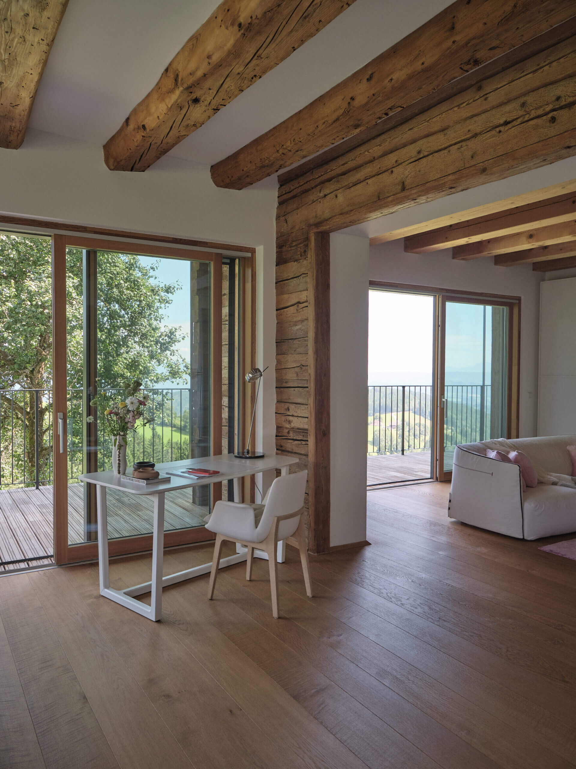 Luxury open-plan suite with a white minimalist desk and rustic wood beam architecture overlooking green hills and valley.